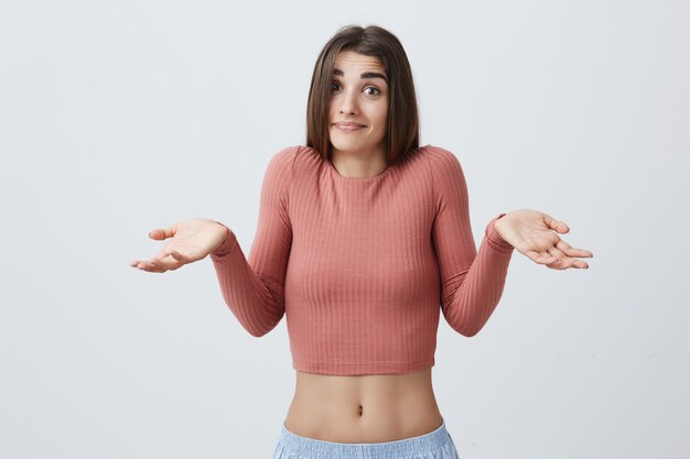 Je n'ai aucune idée. Portrait de jeune séduisante étudiante caucasienne sexy fille aux cheveux longs noirs en chemise rose et short de sport bleu écartant les mains