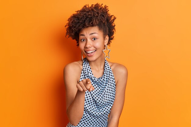 Je ne choisis que toi. Une femme afro-américaine positive dans des vêtements à la mode pointe l'index directement sur vous invite quelqu'un pour la fête a une coiffure élégante isolée sur un fond orange vif.