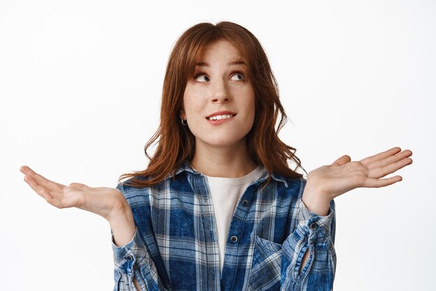 Je ne sais pas Fille rousse confuse haussant les épaules et levant les yeux avec un visage indécis incertain ne peut pas prendre de décision n'a aucune idée debout perplexe sur fond blanc