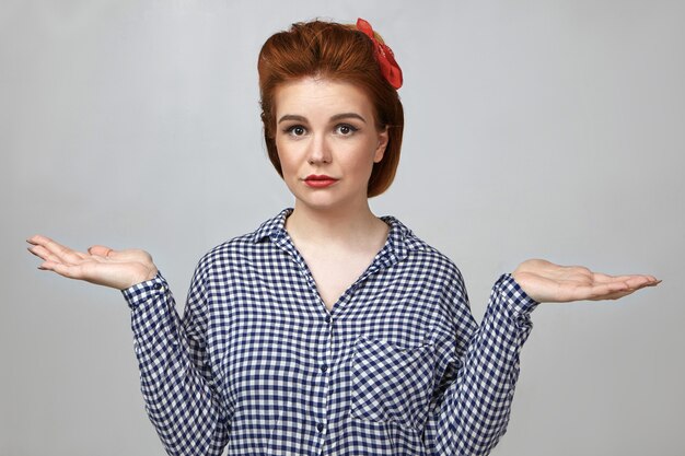 Je ne sais pas. Portrait de femme de 25 ans incertain désemparé portant rouge à lèvres et chemise à carreaux gesticulant avec paumes ouvertes en studio, haussant les épaules