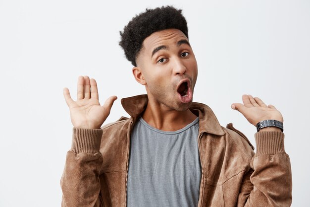 Je suis hors de tout ça. Portrait de jeune homme africain drôle à la peau sombre avec des cheveux bouclés en t-shirt gris sous une veste brune étant choqué d'entendre des nouvelles provocantes sur un vieil ami.