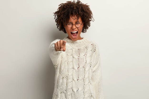 Je te blâme! Une femme afro-américaine stressante avec des cheveux touffus et croustillants pointe l'index directement, crie furieusement, exprime son agacement, se tient au-dessus d'un mur blanc, dit que vous êtes coupable