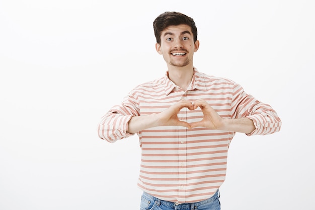 Je Te Donne Mon Cœur. Tir Intérieur De Charmant Petit Ami Européen Avec Moustache, Montrant Le Geste Du Cœur Près De Chet Et Souriant Largement, Avouant Son Amour Et Sa Passion