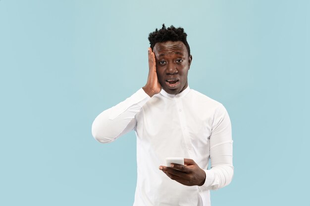 Je viens de perdre un pari. Jeune homme afro-américain avec smartphone isolé sur fond bleu studio, expression faciale. Beau portrait d'homme demi-longueur. Concept d'émotions humaines, expression faciale.