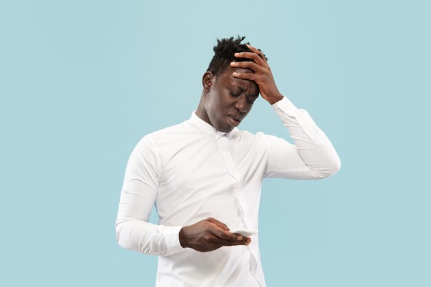 Je viens de perdre un pari. Jeune homme afro-américain avec smartphone isolé sur studio bleu, expression faciale.