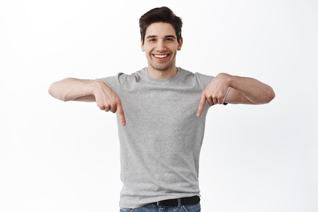 Jetez un oeil Souriant bel homme pointant vers le bas et regardant un magasin de recommandation affirmé ou une offre promotionnelle montrant un espace de copie pour votre logo debout sur fond blanc