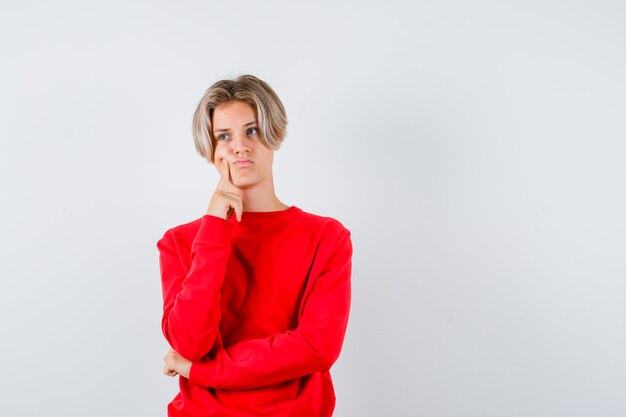 Jeune adolescent en appuyant sur le doigt sur la joue, regardant loin en pull rouge et l'air triste, vue de face.