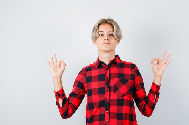 Jeune adolescent en chemise à carreaux montrant un geste de yoga et l'air détendu, vue de face.