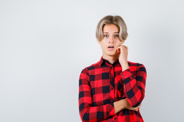 Jeune adolescent en chemise à carreaux touchant la mâchoire et ayant l'air confus, vue de face.