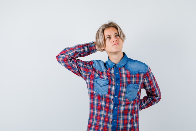 Jeune adolescent gardant la main derrière la tête, levant les yeux en chemise à carreaux et l'air pensif. vue de face.