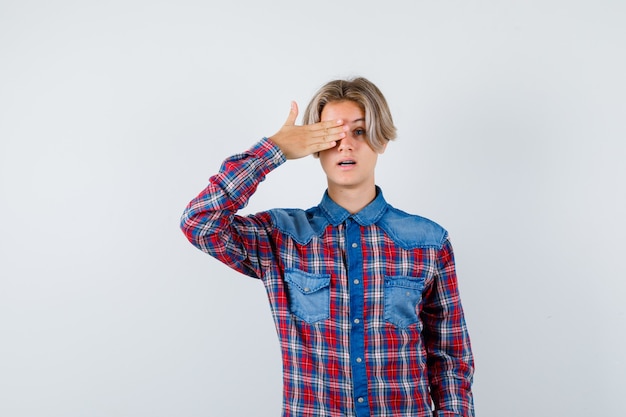 Jeune adolescent avec la main sur l'œil en chemise à carreaux et l'air étonné. vue de face.