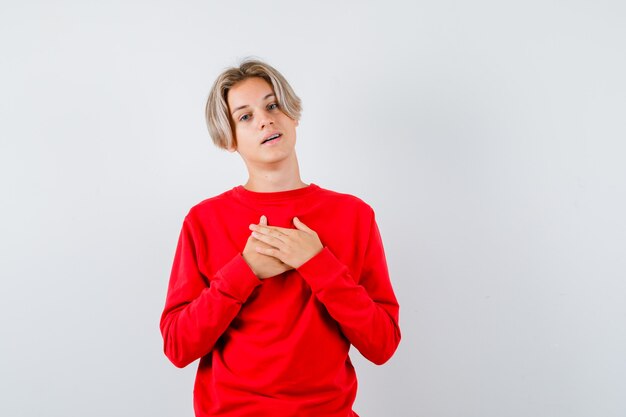 Jeune adolescent avec les mains sur la poitrine en pull rouge et l'air reconnaissant, vue de face.