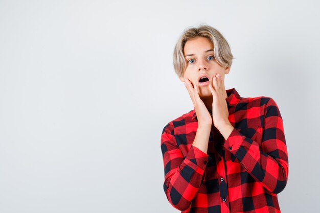 Jeune adolescent avec les mains près de la bouche en chemise à carreaux et l'air choqué. vue de face.