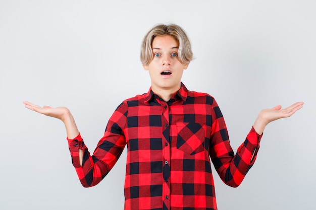 Jeune adolescent montrant un geste impuissant en chemise à carreaux et l'air étonné, vue de face.