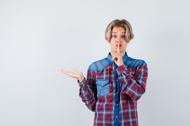 Jeune adolescent montrant un geste de silence, écartant la paume en chemise à carreaux et l'air anxieux, vue de face.