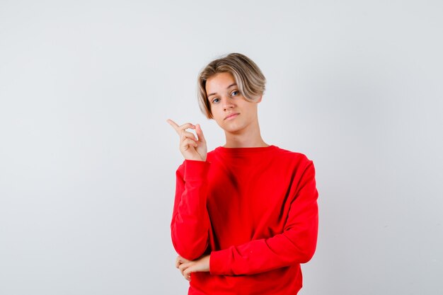 Jeune adolescent pointant vers le coin supérieur gauche en pull rouge et l'air intelligent. vue de face.