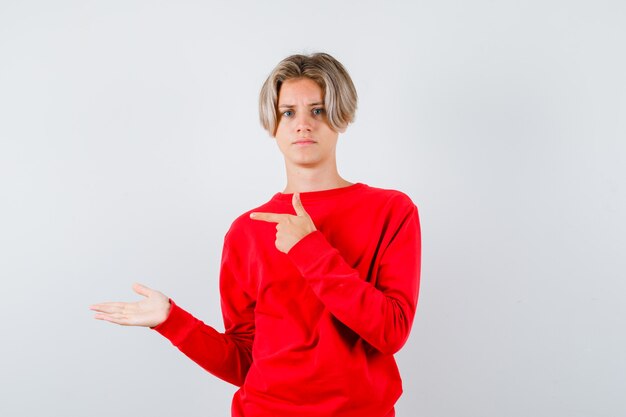 Jeune adolescent pointant vers la gauche, écartant la paume en pull rouge et semblant sérieux, vue de face.