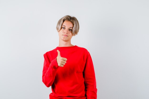 Jeune adolescent en pull rouge montrant le pouce vers le haut et l'air satisfait, vue de face.