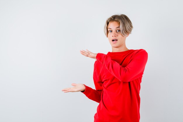 Jeune adolescent en pull rouge montrant un signe de taille et à la perplexité, vue de face.