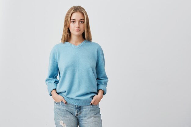 Jeune adolescente aux cheveux blonds tendre avec une peau saine portant un haut bleu à la recherche avec une expression sérieuse ou pensive. Modèle femme caucasienne avec les mains dans les poches posant à l'intérieur