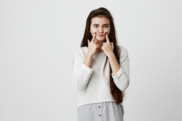 jeune adolescente brune bouleversée faisant un faux sourire avec ses doigts étirant les coins de sa bouche. Portrait de femme essayant de rester positif