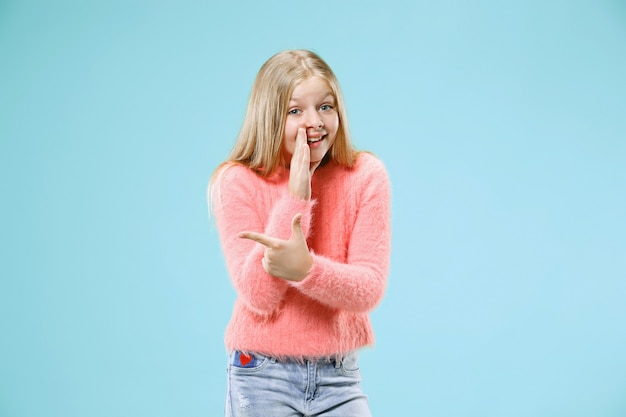 Jeune adolescente chuchotant un secret derrière sa main isolé sur un mur de studio bleu à la mode