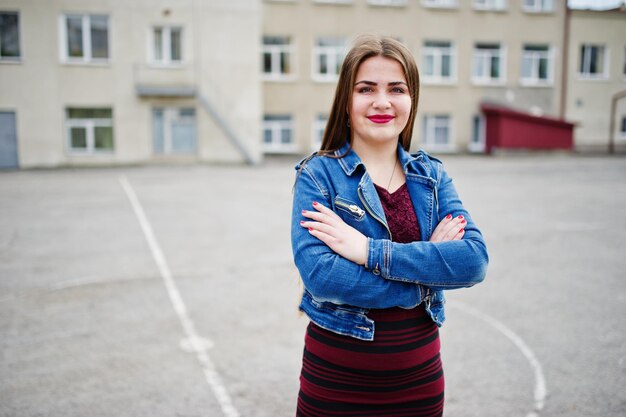 Photo gratuite jeune adolescente potelée portant une robe rouge et une veste en jean posée contre l'arrière-cour de l'école