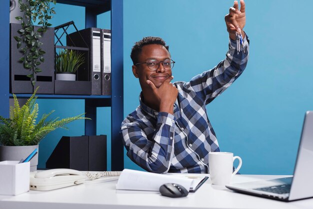 Jeune adulte propriétaire d'une maison tenant les clés d'une nouvelle maison achetée pendant son mandat. Homme confiant et heureux ayant les clés de l'investissement immobilier récent tout en étant assis au bureau dans un espace de travail moderne.