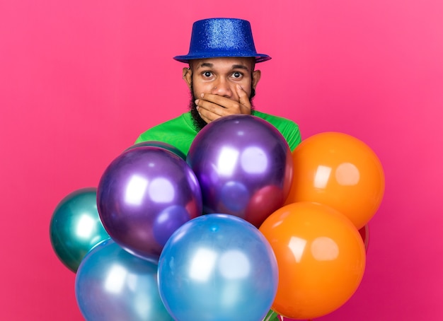 Photo gratuite un jeune afro-américain effrayé portant un chapeau de fête debout derrière des ballons a couvert la bouche avec la main