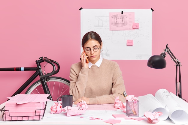 Une jeune architecte talentueuse fait des croquis de la planification du bâtiment a une conversation téléphonique impliquée dans le processus de travail entourée de papiers plans poses sur le bureau