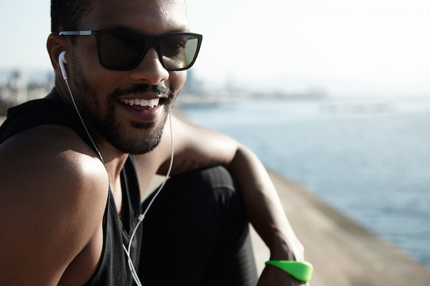 Jeune athlète africain charismatique portant des nuances à la mode et une tenue noire, regardant heureux et joyeux assis au bord de la mer contre le ciel bleu et la mer