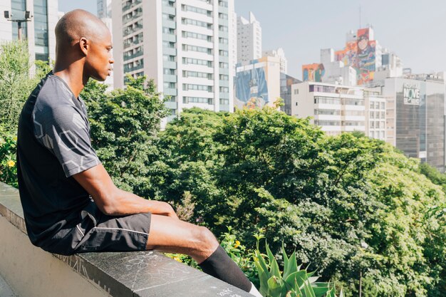 Jeune athlète masculin africain assis sur le bord du toit surplombant la ville