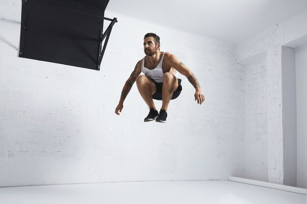 Jeune athlète masculin barbu et tatoué montre des mouvements calisthéniques sauts en hauteur dans l'air, isolé dans la salle blanche du centre de remise en forme