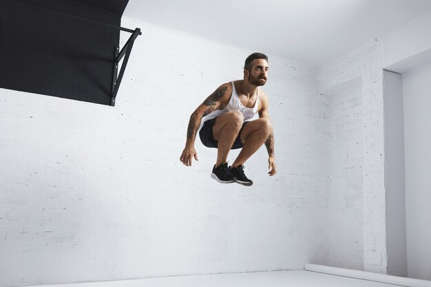 Un jeune athlète masculin barbu et tatoué montre des mouvements de gymnastique, saute haut dans les airs à côté de la barre de traction noire, portant un t-shirt de réservoir blanc, isolé dans la salle blanche du centre de remise en forme