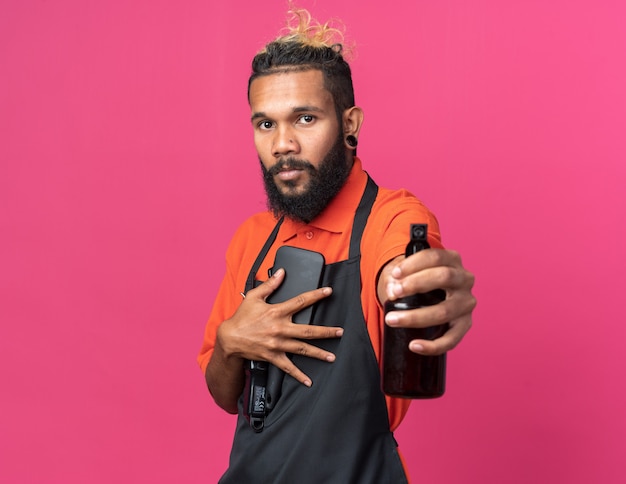 Photo gratuite jeune barbier afro-américain confiant en uniforme debout en vue de profil tenant des outils de barbier regardant à l'avant s'étirant la laque pour cheveux vers l'avant