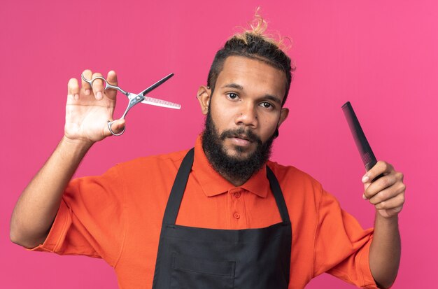 Jeune barbier afro-américain confiant en uniforme tenant des ciseaux et un peigne