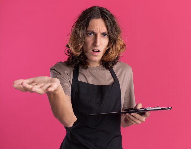 Jeune barbier beau désemparé portant l'uniforme tenant le presse-papiers montrant la main vide