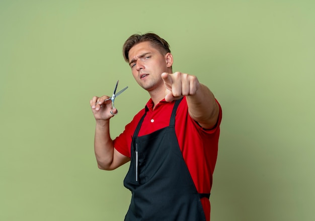 Jeune barbier blond confiant dans des points uniformes à la caméra et détient des ciseaux isolés sur l'espace vert olive avec copie espace