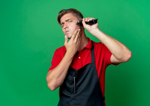 Jeune barbier blond confiant en uniforme tient la barbe de coupe du visage avec tondeuse à cheveux isolé sur espace vert avec espace copie