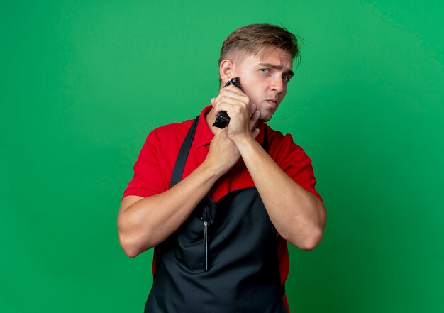 Jeune barbier blond confiant en uniforme tient la barbe de coupe du visage avec tondeuse à cheveux isolé sur espace vert avec espace copie