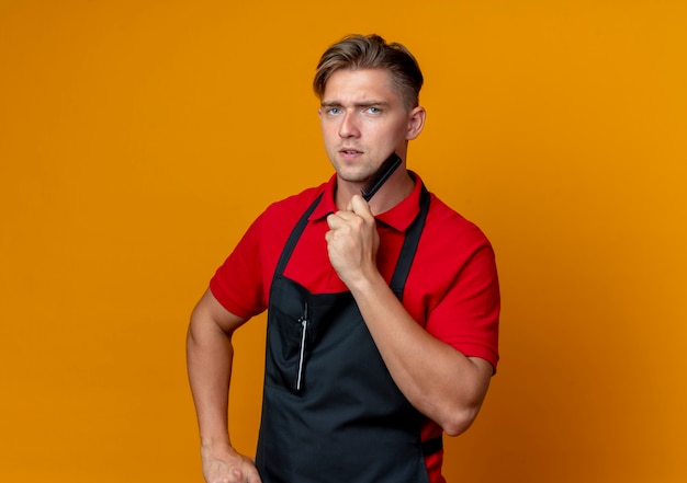 Jeune barbier blond confiant en uniforme tient et met le peigne sur le menton isolé sur l'espace orange avec copie espace