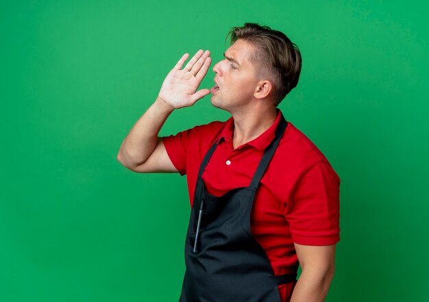 Jeune barbier blond sérieux en uniforme regarde le côté fait semblant d'appeler quelqu'un tenant la main près de la bouche isolé sur un espace vert avec copie espace