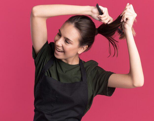 Une jeune barbier brune ravie en uniforme fait semblant de couper les cheveux avec des ciseaux à effiler les cheveux en regardant le côté isolé sur un mur rose avec un espace de copie