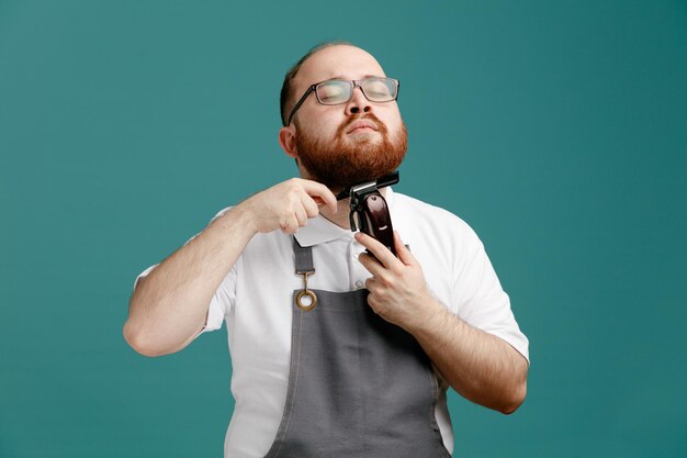 Jeune barbier concentré portant un uniforme et des lunettes tenant un peigne teaser et une tondeuse à cheveux taillant sa propre barbe avec les yeux fermés isolés sur fond bleu