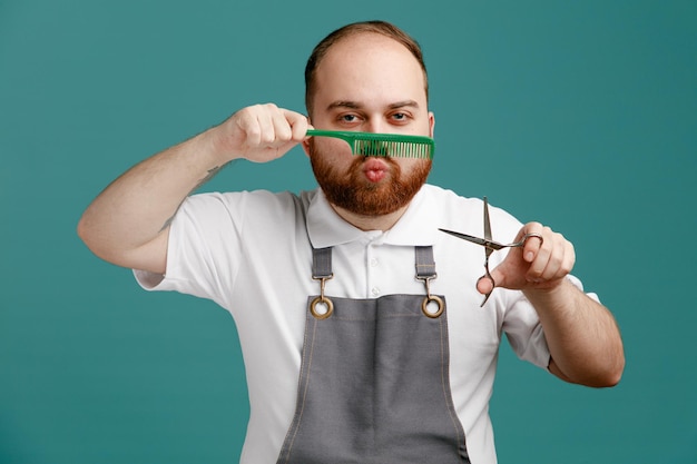 Jeune barbier espiègle portant une chemise blanche et un tablier de coiffeur tenant des ciseaux regardant la caméra faisant un geste de moustache avec un peigne isolé sur fond bleu