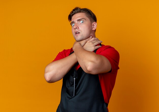 Jeune barbier homme blond effrayé en uniforme fait semblant de s'étouffer avec les mains isolées sur l'espace orange avec copie espace