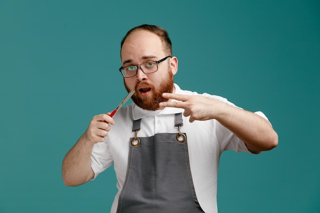 Jeune barbier impressionné portant l'uniforme et des lunettes montrant un rasoir droit regardant la caméra montrant un signe de paix isolé sur fond bleu