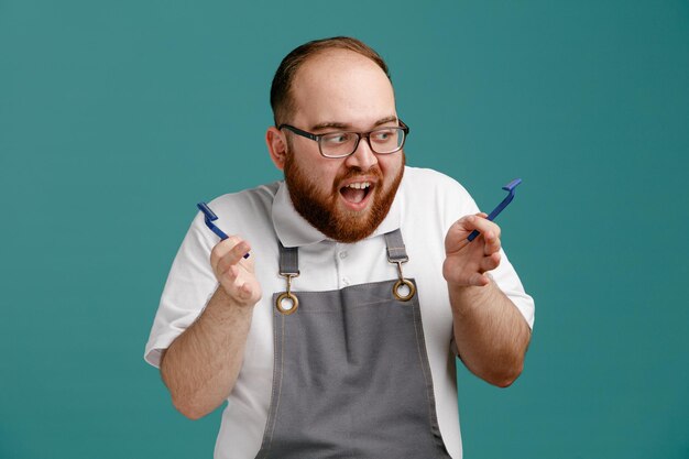 Jeune barbier impressionné en uniforme et lunettes tenant des rasoirs en regardant l'un d'eux isolé sur fond bleu