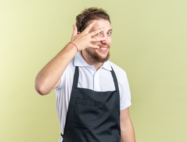 Jeune barbier masculin joyeux portant le visage couvert d'uniforme avec des mains d'isolement sur le mur vert olive