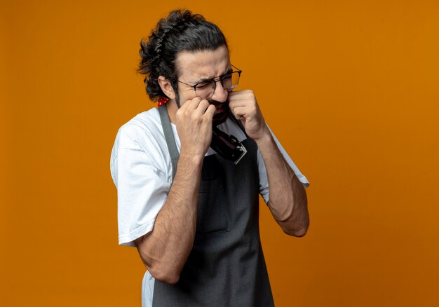 Jeune barbier de race blanche en uniforme portant des lunettes tenant une tondeuse à cheveux mettant les mains sur les joues souffrant de maux de dents avec les yeux fermés